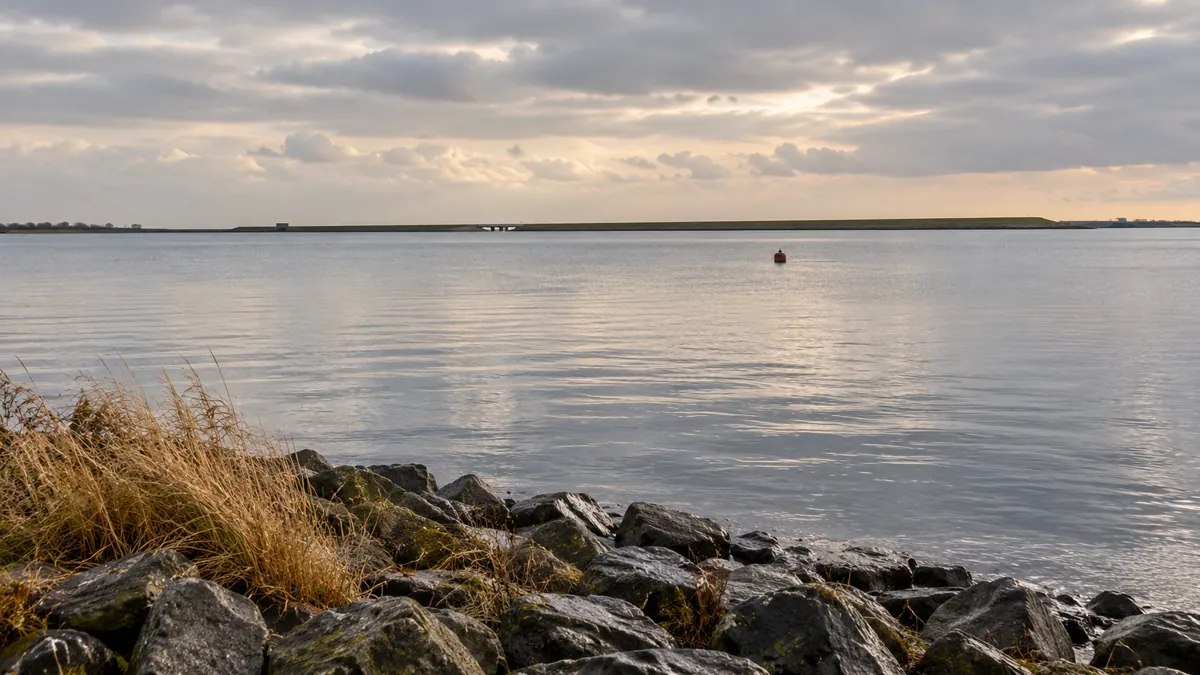 Rustig water langs een stenen oever, met een lage dam aan de horizon.