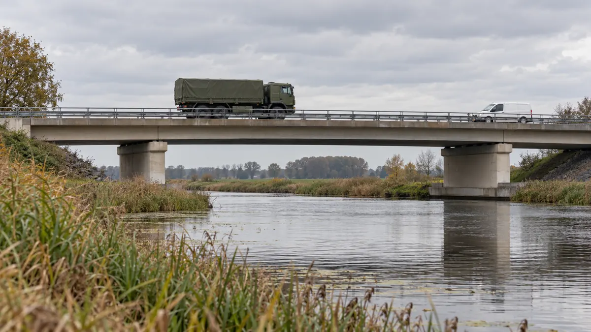 Een militaire vrachtwagen en een bestelbus op een betonnen brug in een vlak landschap.