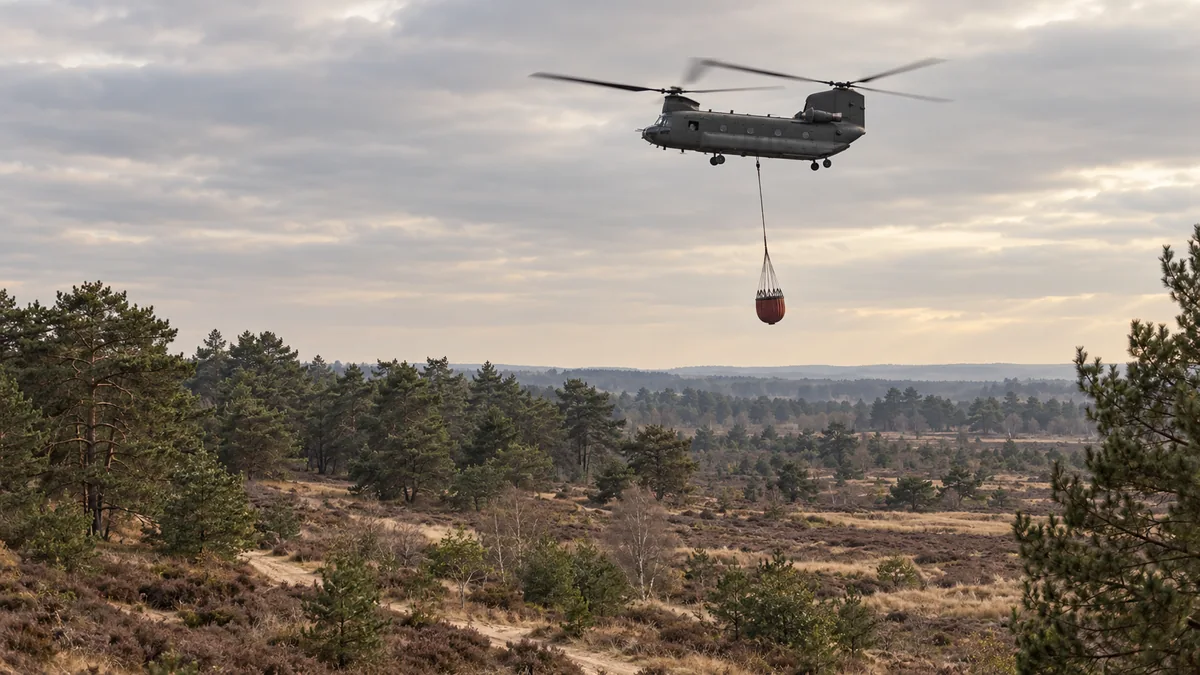 Een transporthelikopter met een grote waterzak vliegt boven heide en naaldbos.